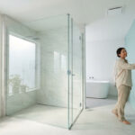 Une femme touche un mur en mosaïque bleu clair dans une salle de bain lumineuse avec douche vitrée et baignoire.